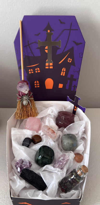photo of actual pieces and complete set bundle Witch’s broom, assorted crystal tumbles, strawberry quartz cauldron, green jasper, labradorite and green jasper cubes, black tourmaline raw stone, wish bottle with dragon’s blood chips, tumbles and pieces of mica. Background shows lid to specialty paper box included. Coffin shaped two piece box, purple and orange decorated with a halloween house, bats and tomb stones