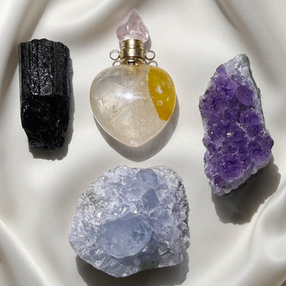 Four raw crystals and a heart-shaped glass perfume bottle arranged on a white textured background.  Black Tourmaline, Golden Healer Heart, Amethyst Geode and Celestite Geode
