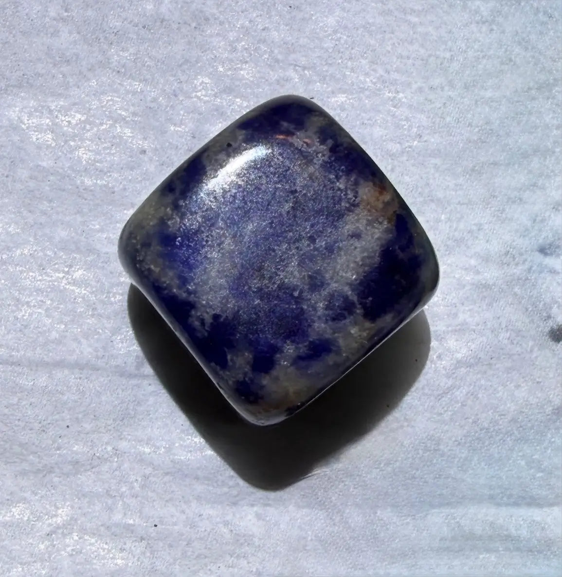 A polished square tumbled gemstone with rounded corners, featuring a deep blue and white mottled pattern, resting on a textured light gray surface.