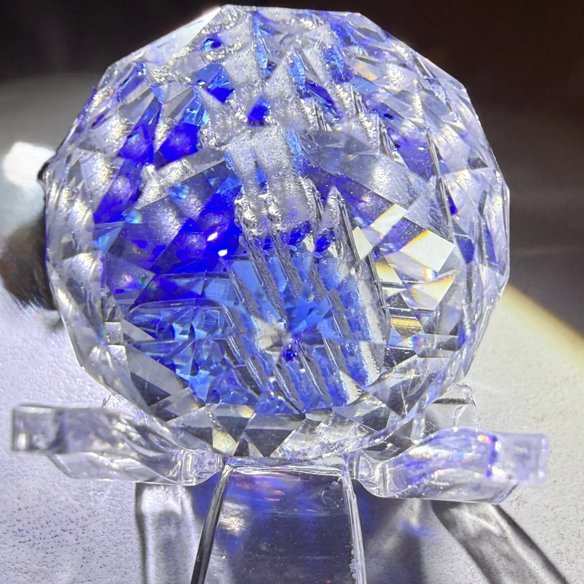 A faceted clear crystal ball sculpture or decorative object displayed on a clear stand, with strong blue reflections and some white highlights from the cut geometric facets.