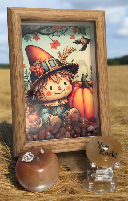 A decorative arrangement featuring a framed autumn-themed illustration of a smiling scarecrow with a pumpkin, surrounded by assorted brownish-red carnelian stones at the bottom. In front of the frame, there is a polished stone apple with a jeweled leaf and a tan stone with the engraved phrase “You Rock!” displayed in a field of hay