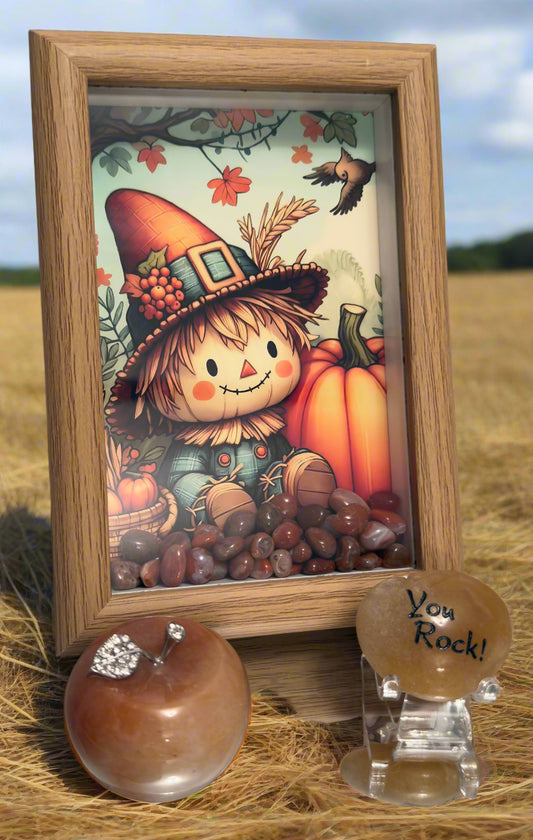 A decorative arrangement featuring a framed autumn-themed illustration of a smiling scarecrow with a pumpkin, surrounded by assorted brownish-red carnelian stones at the bottom. In front of the frame, there is a polished stone apple with a jeweled leaf and a tan stone with the engraved phrase “You Rock!” displayed in a field of hay