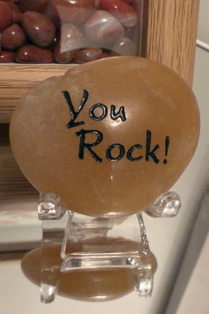 Orange Calcite Inspiration Stone with the engraved phrase “You Rock!” displayed on a clear stand, all placed on a reflective surface. Main Street Crystals and Minerals