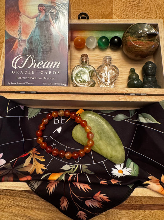 A wooden box containing a deck of "Dream Oracle Cards," colorful round stones, small perfume bottles, jade figures, a gemstone bracelet, and a jade gua sha tool on top of a dark floral patterned scarf.
