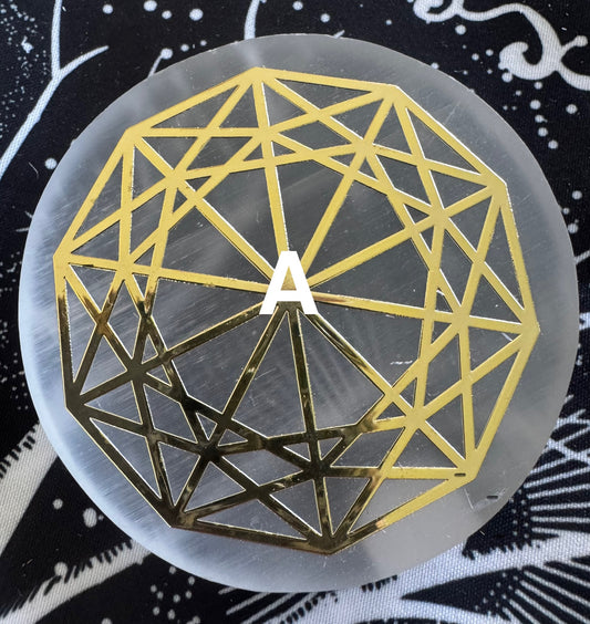 A round, white selenite plate with a gold geometric design and the letter "A" in the center on a black and white patterned background.