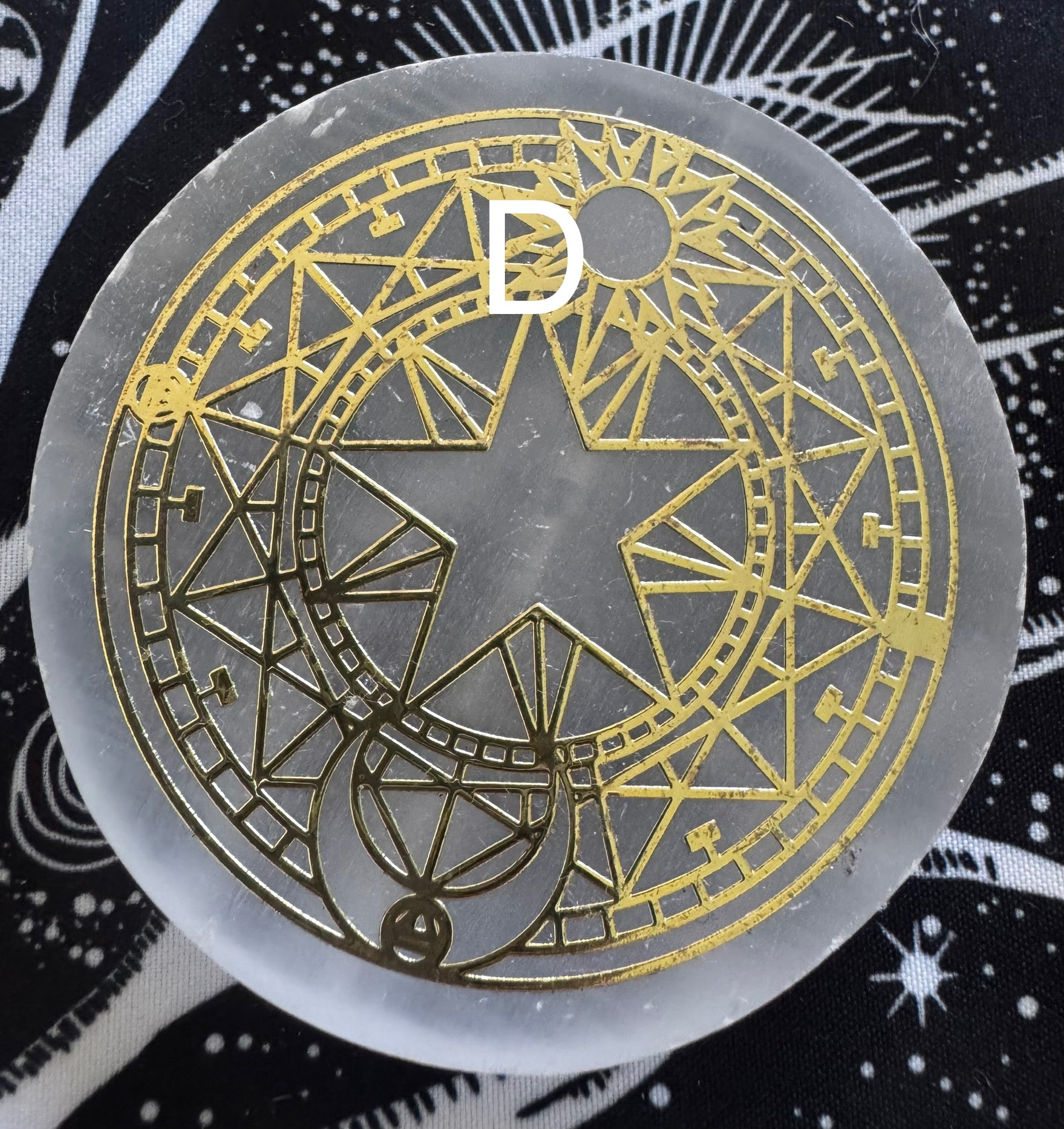 Close-up of a round, translucent selenite plate with a gold geometric design featuring a star, sun, and moon, overlaid with a white letter "D", against a black and white patterned fabric background.
