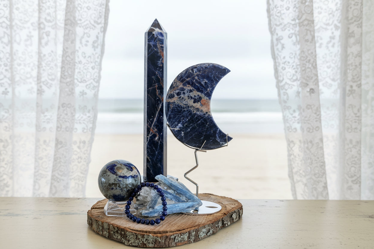 Large Sunset Sodalite Tower, Crescent Moon, Lapis Sphere and Bracelet with Blue Kyanite Raw Specimen sitting on a wood plank in front of a window with sheer curtains and a muted beach scene behind