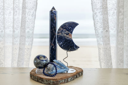 Large Sunset Sodalite Tower, Crescent Moon, Lapis Sphere and Bracelet with Blue Kyanite Raw Specimen sitting on a wood plank in front of a window with sheer curtains and a muted beach scene behind