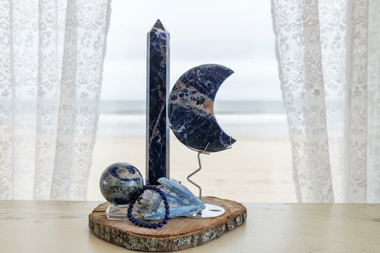Large Sunset Sodalite Tower, Crescent Moon, Lapis Sphere and Bracelet with Blue Kyanite Raw Specimen sitting on a wood plank in front of a window with sheer curtains and a muted beach scene behind