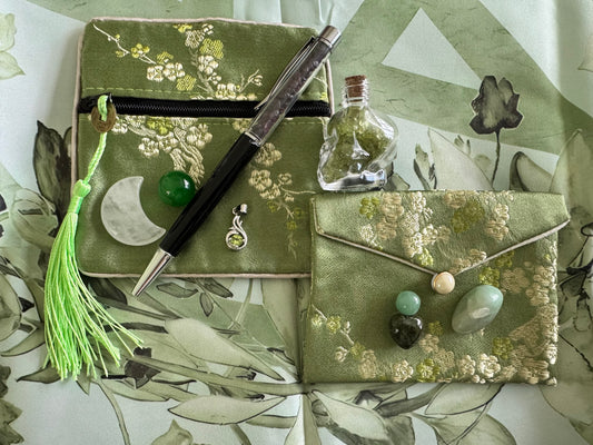 Product shot of two green floral pouches, a black and silver pen, gemstones, a skull-shaped bottle filled with green stones, and a green tassel on a floral patterned surface.
