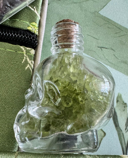Skull-shaped glass bottle filled with green crystals, sealed with a cork stopper.
