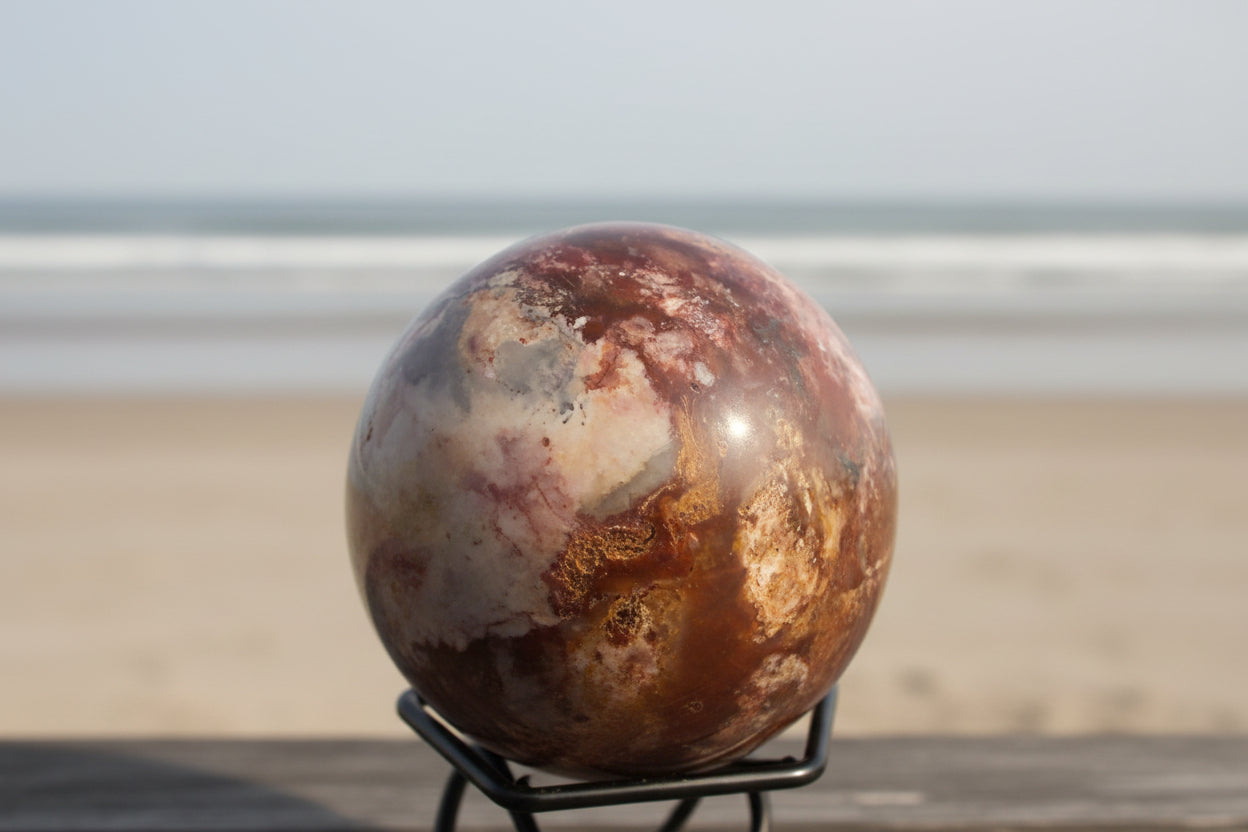 Polychrome jasper sphere in a small stand on the beach Colors are bright oranges, blue, ivory and white
