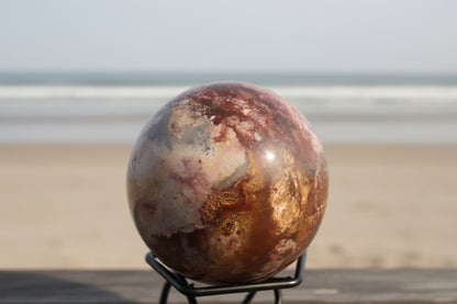 Polychrome jasper sphere in a small stand on the beach Colors are bright oranges, blue, ivory and white
