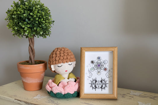 Crocheted doll with brown hair and pink flower, next to a framed artwork on a wooden surface.