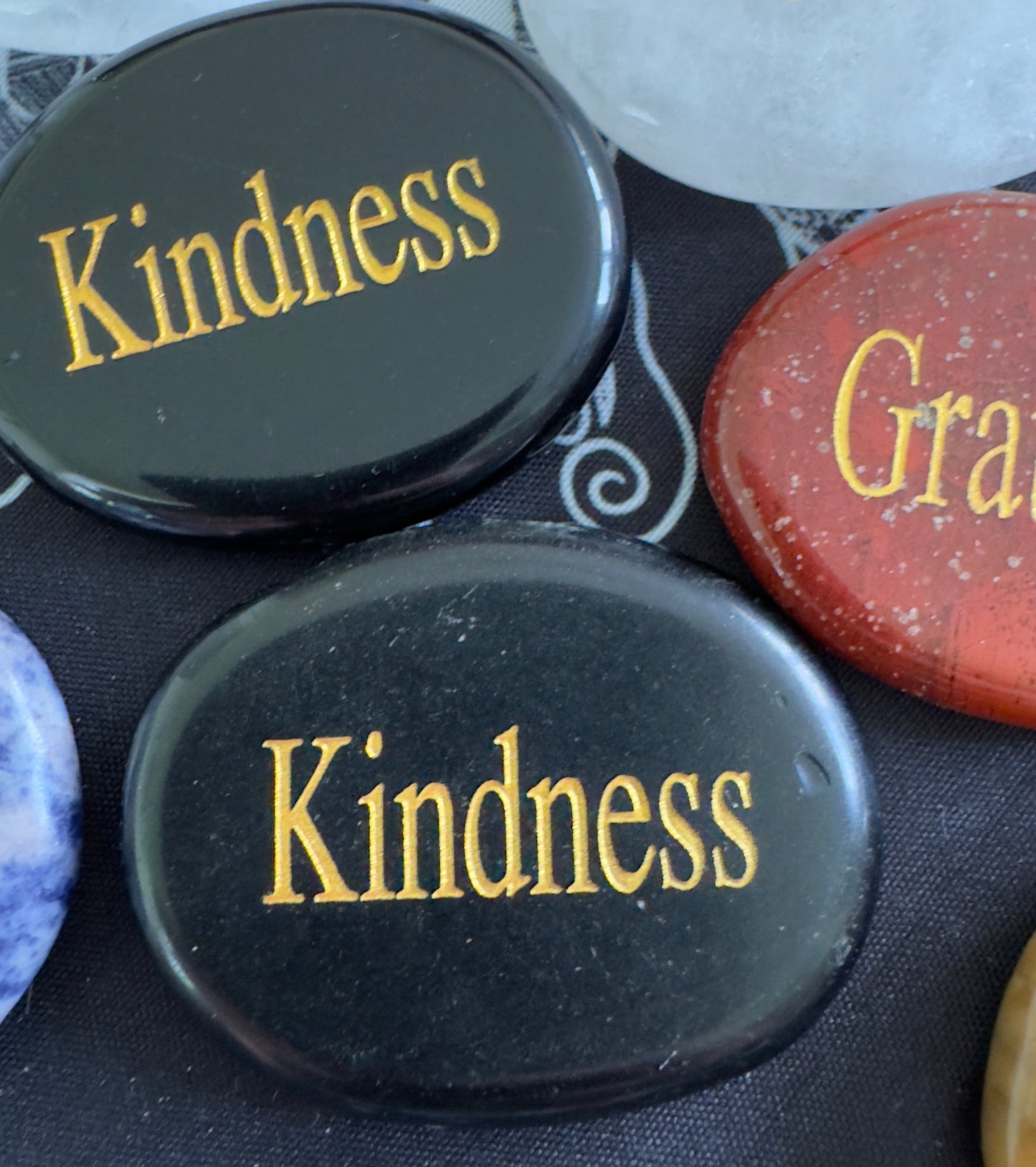 Close-up of several polished stones with gold lettering. The stones are black, red, white, and blue. Some say "Kindness", and another says "Gratitude".
