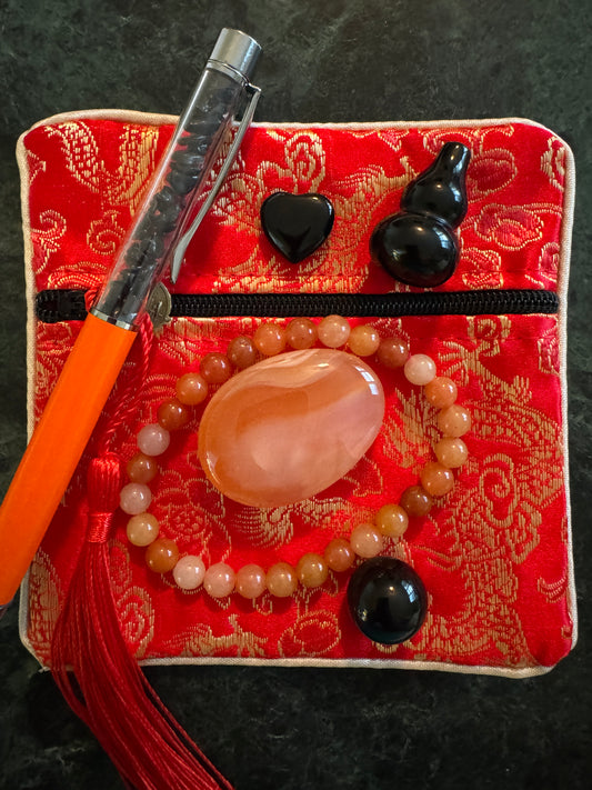 Overhead view of a red brocade zippered bag with a pen, black obsidian heart, black obsidian gourd, agate bracelet, and an agate palm stone.