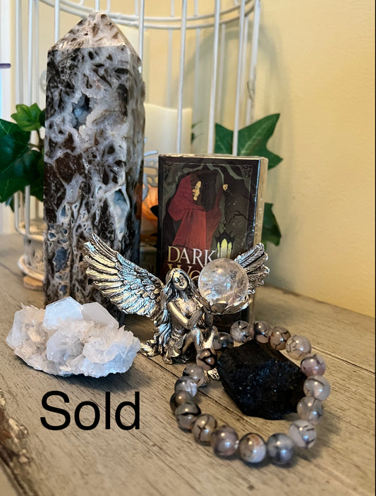 A crystal tower, quartz cluster, angel figurine holding a clear sphere, and bracelet with black and marbled gray beads on a wooden surface. The words "Sold" is imposed in front.