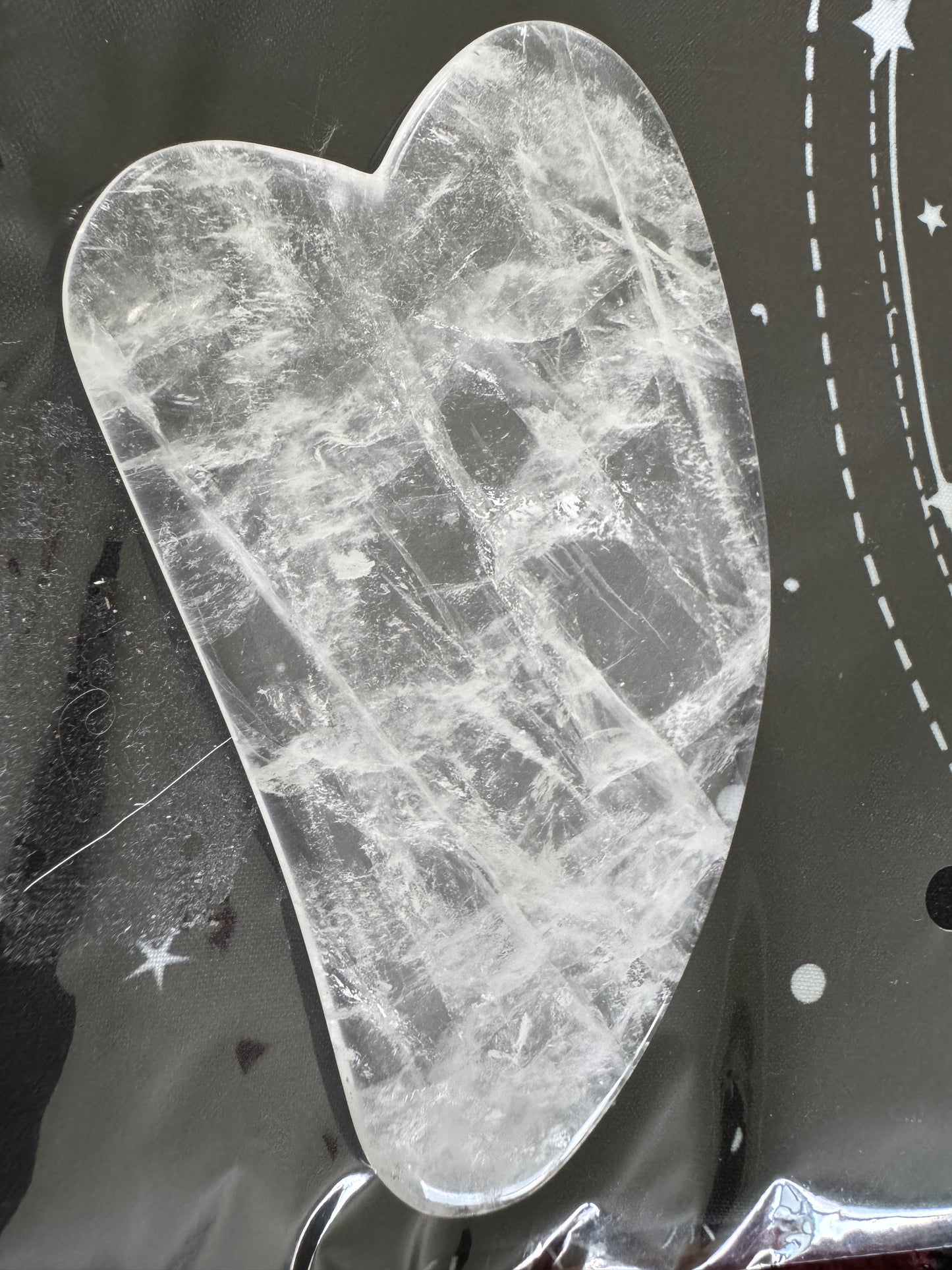 Heart-shaped clear quartz gua sha tool on a black surface with white celestial designs.
