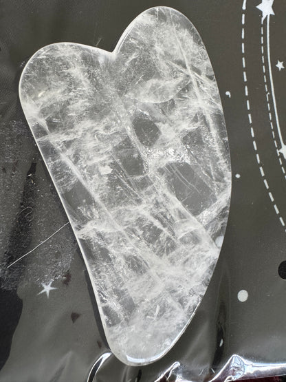 Heart-shaped clear quartz gua sha tool on a black surface with white celestial designs.

