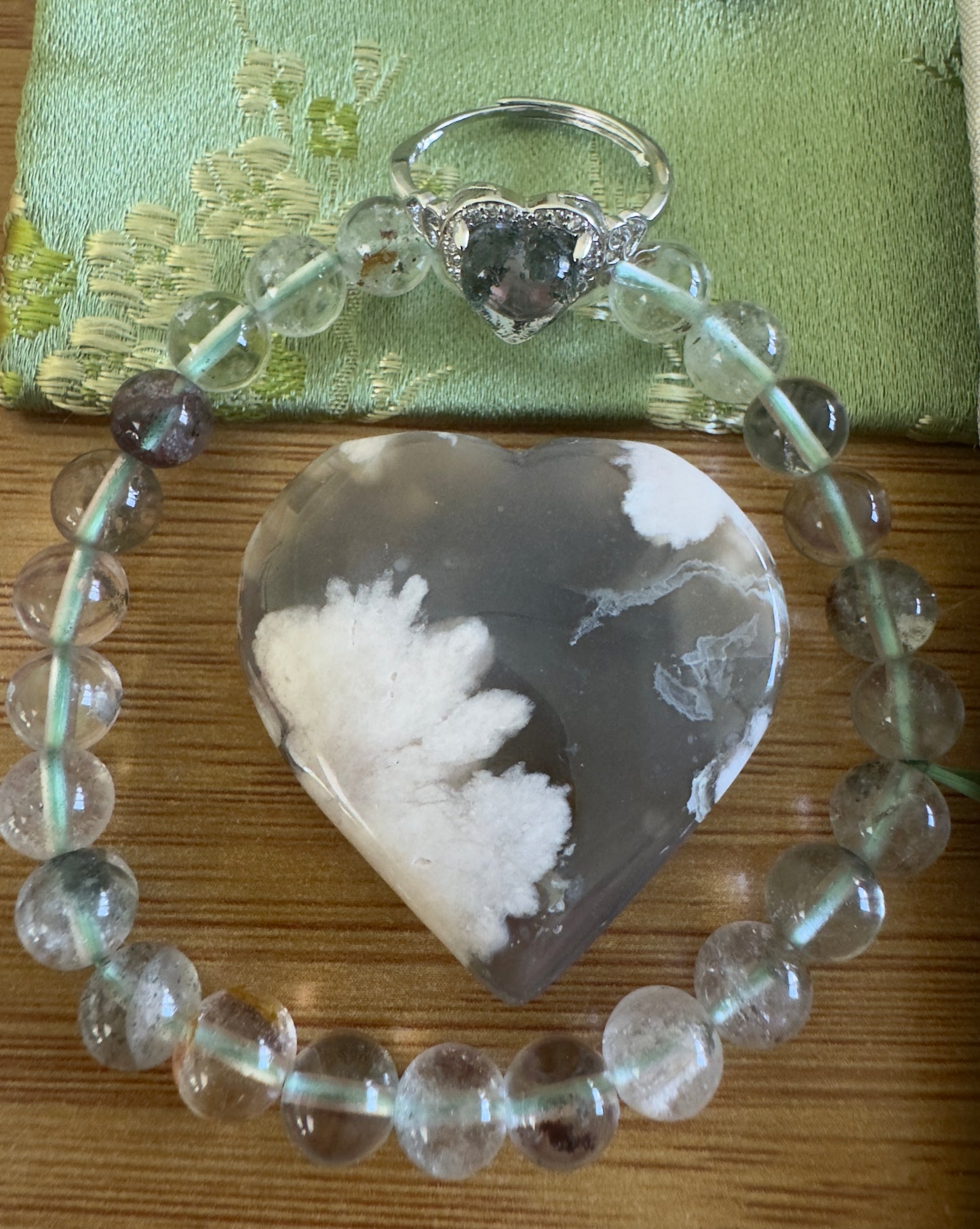 A heart-shaped agate pendant, a beaded bracelet, and a heart-shaped gemstone ring on a wooden surface with a green fabric backdrop.