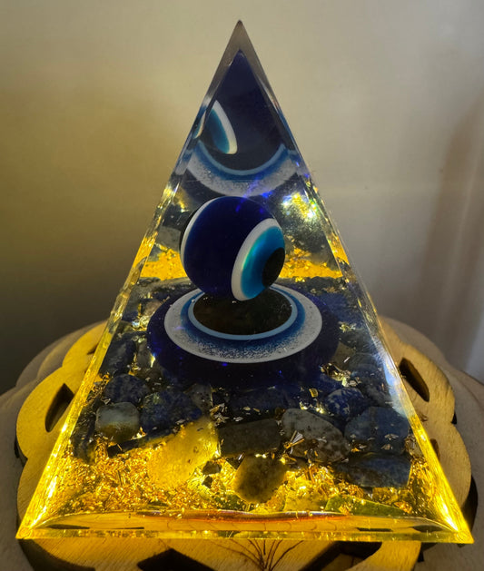 Close-up of a pyramid-shaped resin sculpture, illuminated with warm light, featuring a prominent blue Evil Eye bead and embedded gold flakes and lapis lazuli stones.