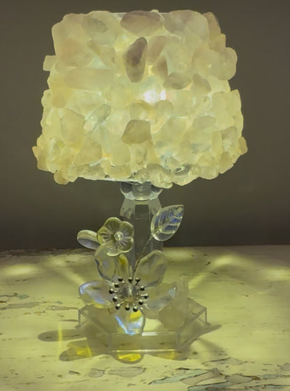 Illuminated lamp featuring a shade covered in translucent, yellowish stones, atop a clear, floral-themed base.