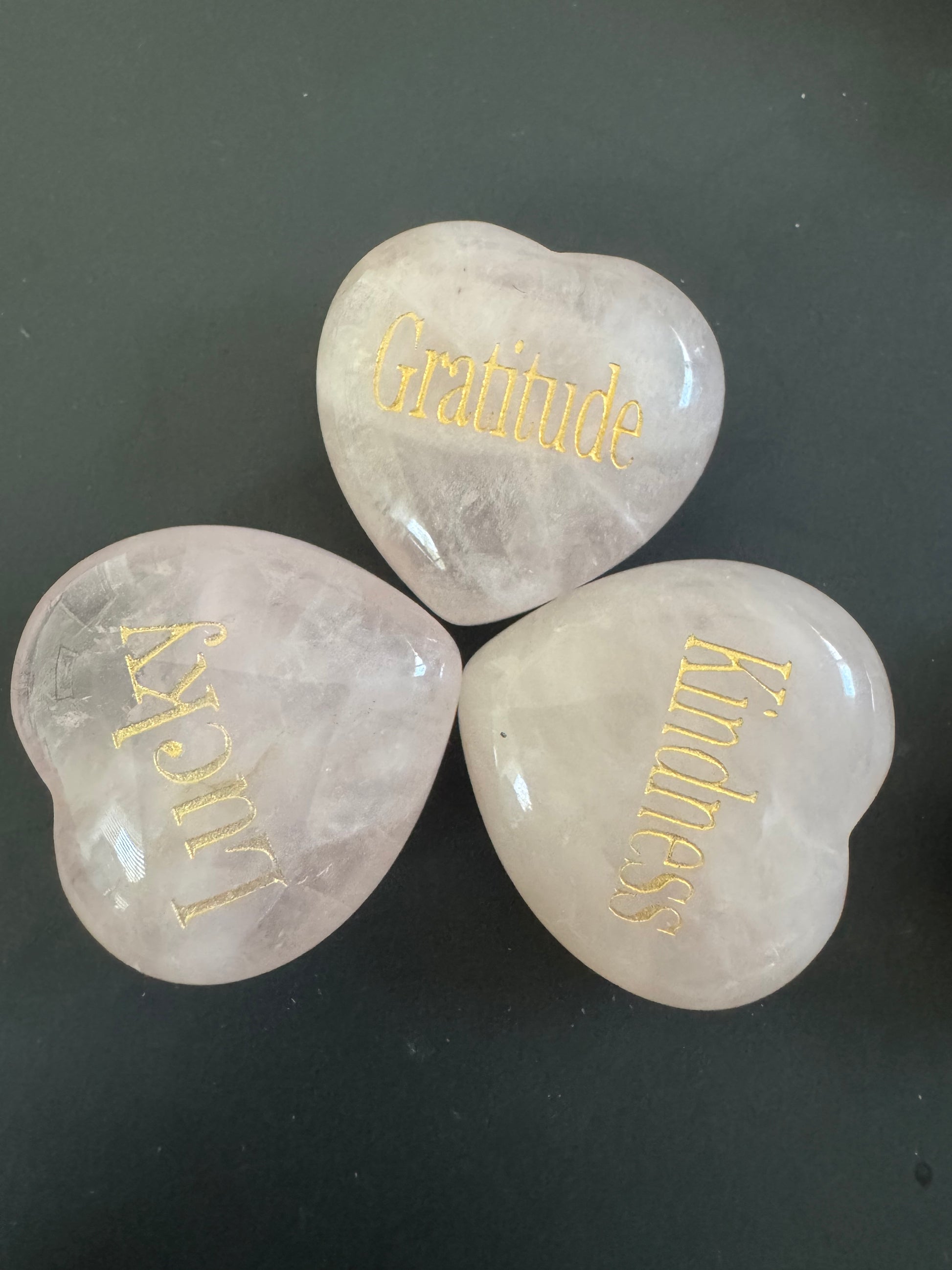 Three heart-shaped rose quartz stones with gold lettering: "Gratitude", "Lucky", and "Kindness" on a dark background.
