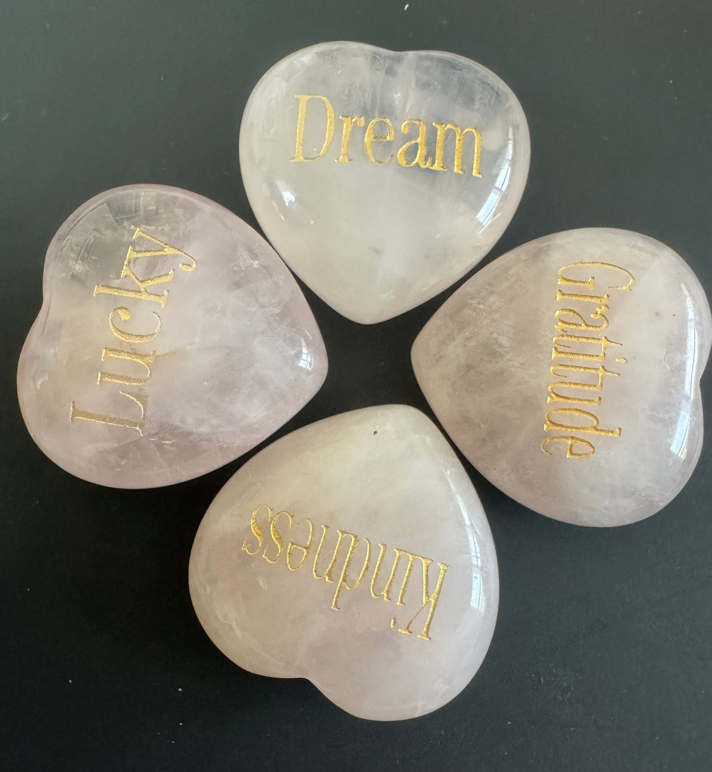 Four heart-shaped, rose quartz stones with gold lettering spelling out "Dream," "Lucky," "Gratitude," and "Kindness."