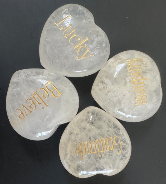 Four heart-shaped clear stones with gold lettering that says, "Lucky", "Kindness", "Believe", and "Gratitude" arranged in a cloverleaf pattern on a dark gray surface.