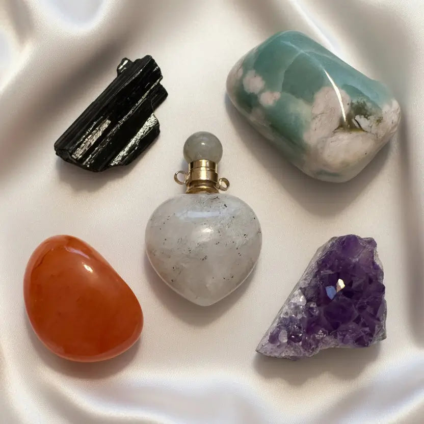 Close up photo of five crystals, moonstone pendant, amethyst cluster, flower agate palm stone, black tourmaline raw specimen and carnelian tumble.  All on a soft off white fabric
