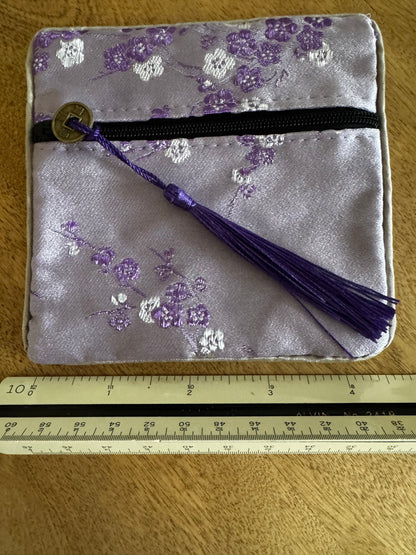 Small, square, lilac satin pouch with plum blossom pattern, black zipper, purple tassel, and gold Chinese coin pull, next to a white architect's scale.