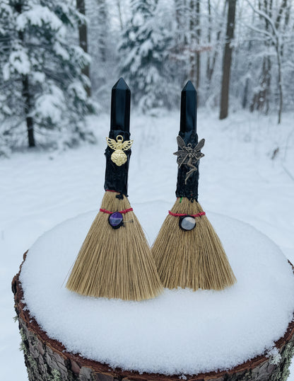 two hand crafted besoms with black obsidian finials  angel charm and fairy charm both brooms placed standing on a snowy tree stump with snow forest background