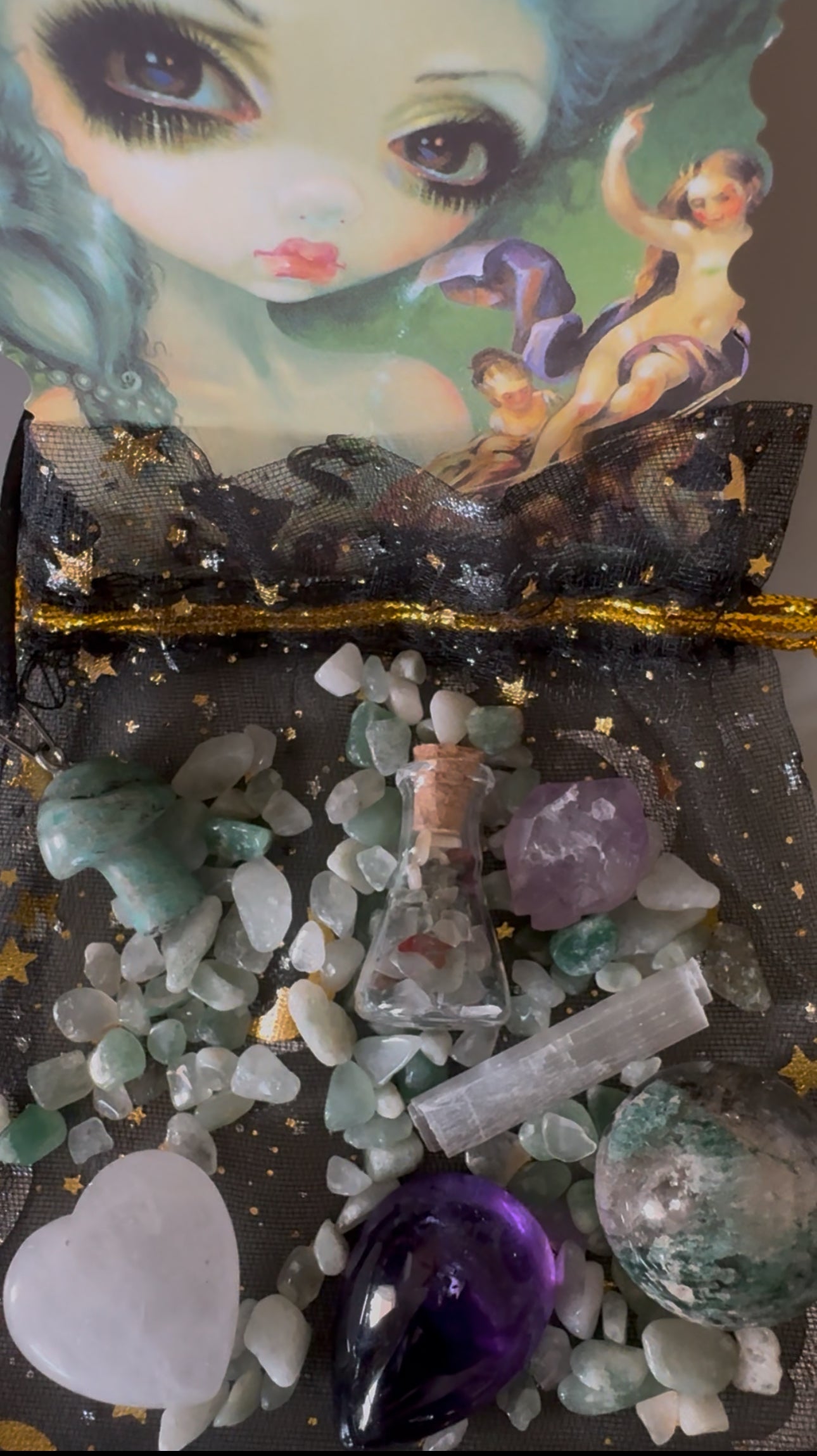 A black drawstring bag with gold stars and a gold ribbon is filled with various crystals, including green aventurine, amethyst, and selenite. A small glass bottle with a cork stopper is also in the bag, along with a whimsical doll print as a backdrop.
