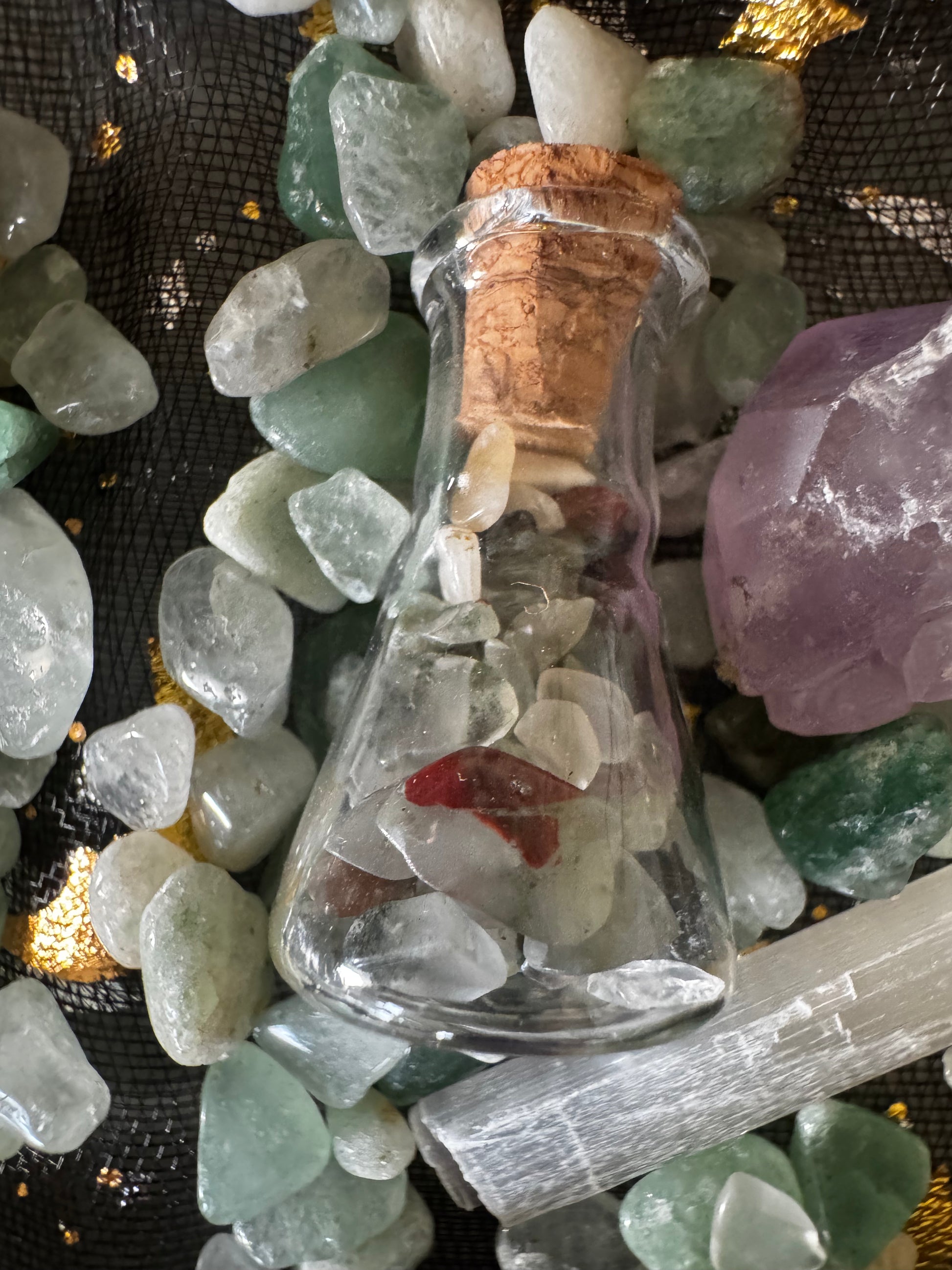 Close-up of a corked glass bottle filled with white and red stones, surrounded by green and white polished stones, a chunk of purple amethyst, and a white selenite stick on a black and gold cloth background.