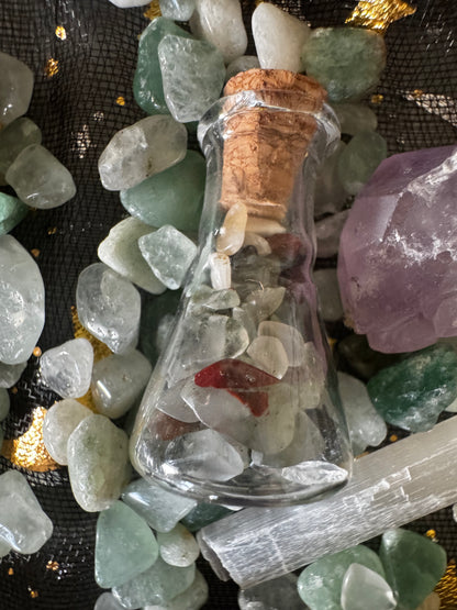 Close-up of a corked glass bottle filled with white and red stones, surrounded by green and white polished stones, a chunk of purple amethyst, and a white selenite stick on a black and gold cloth background.