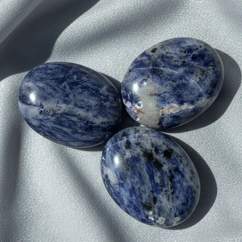pictured three sodalite palm stones with silver fabric behind soft blues, some black and then off white colors