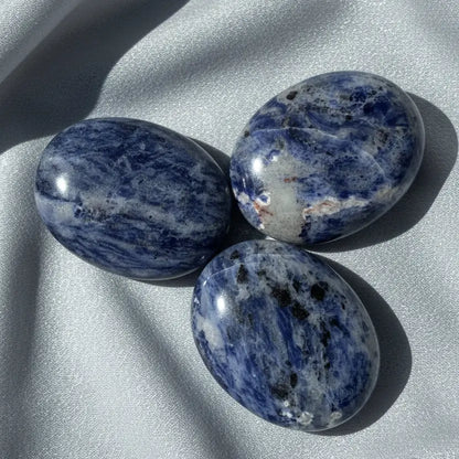pictured three sodalite palm stones with silver fabric behind soft blues, some black and then off white colors
