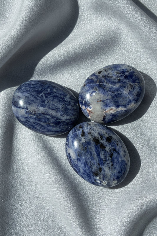 pictured three sodalite palm stones with silver fabric behind soft blues, some black and then off white colors