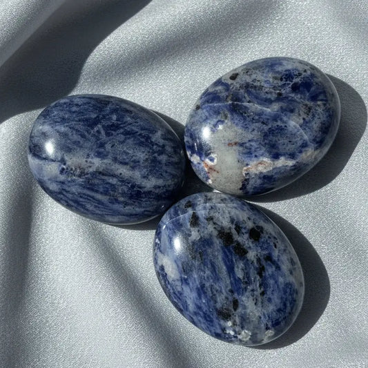 pictured three sodalite palm stones with silver fabric behind soft blues, some black and then off white colors