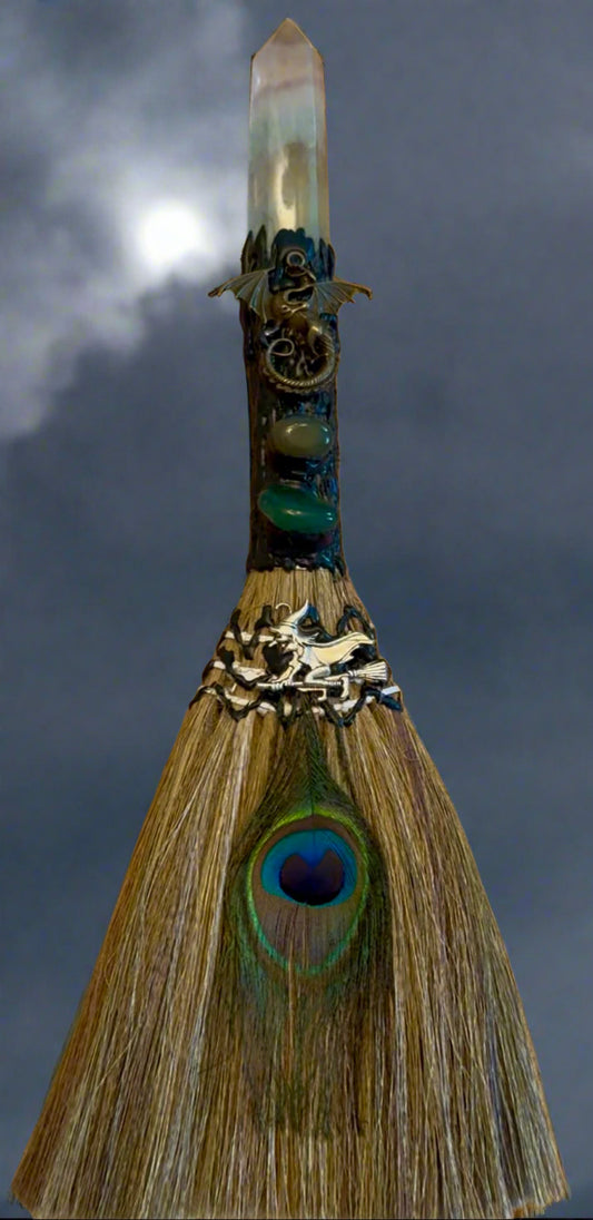 A decorated broom with a quartz crystal top, dragon figurines, gemstones, a peacock feather, and a witch icon.