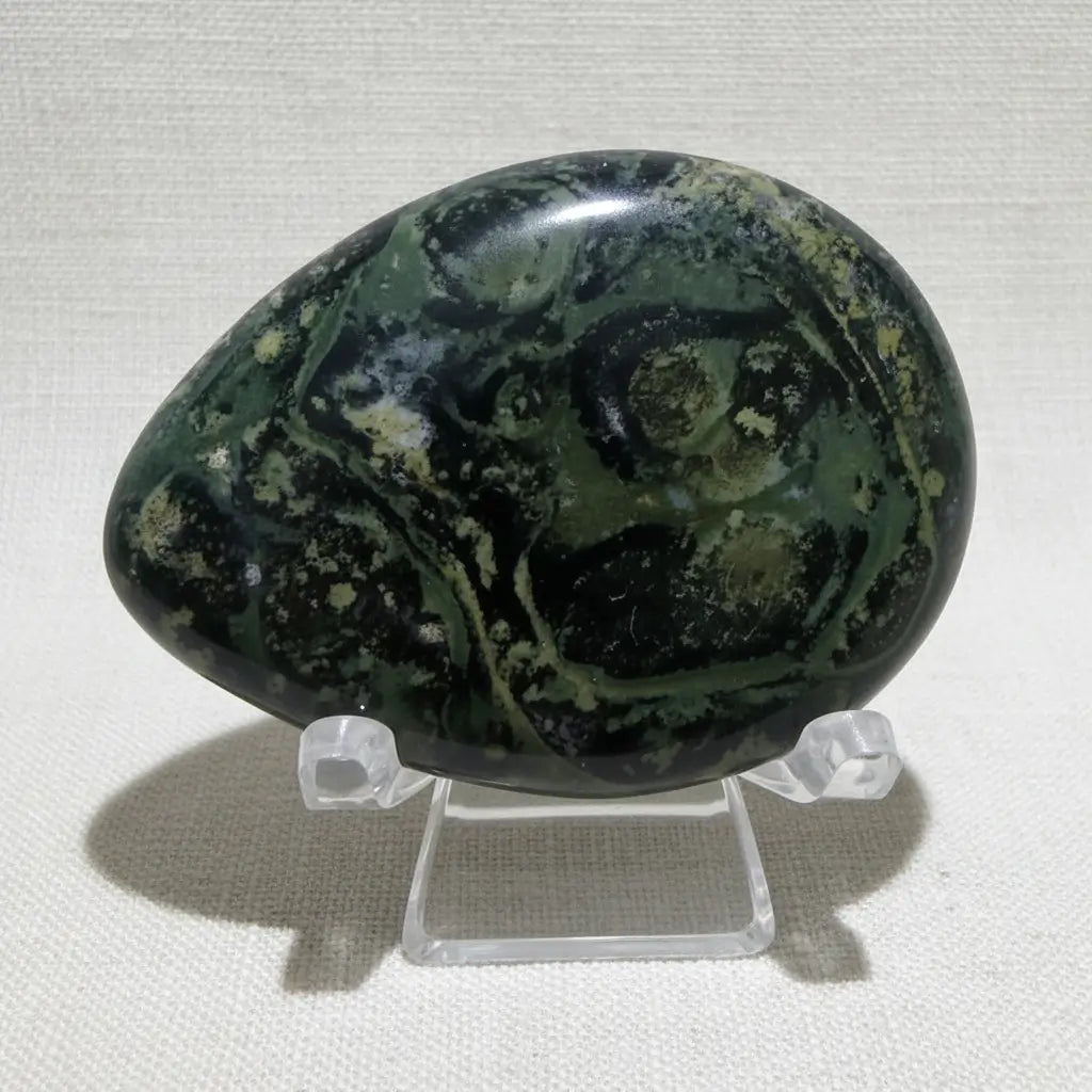 close up of beautiful green with black kambaba jasper worry stone on a clear stand 