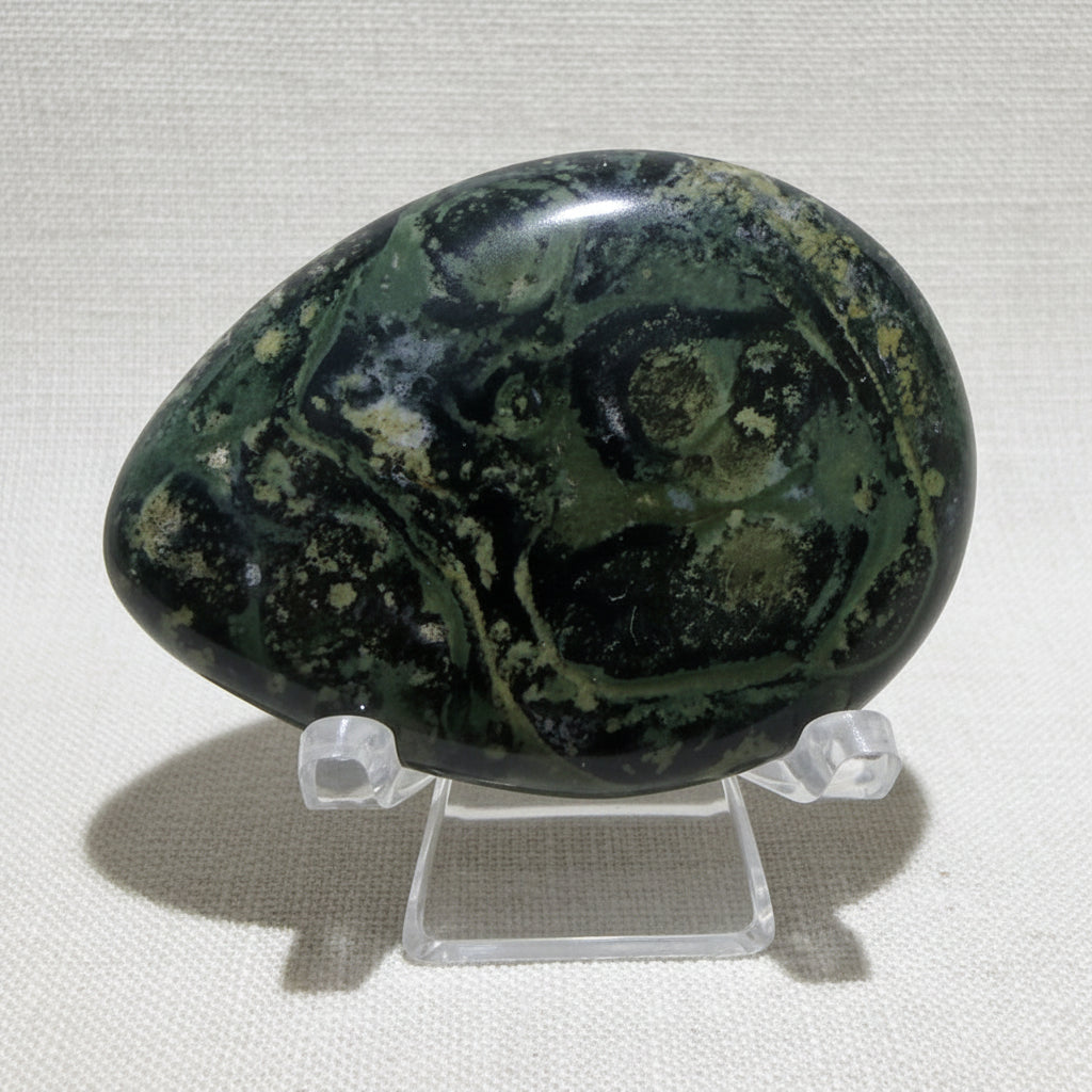 close up of beautiful green with black kambaba jasper worry stone on a clear stand 