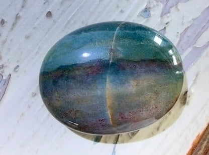 A polished oval cabochon ocean jasper palm stone with a marbled pattern of teal, blue, purple, and brown hues, resting on a distressed white wooden surface.