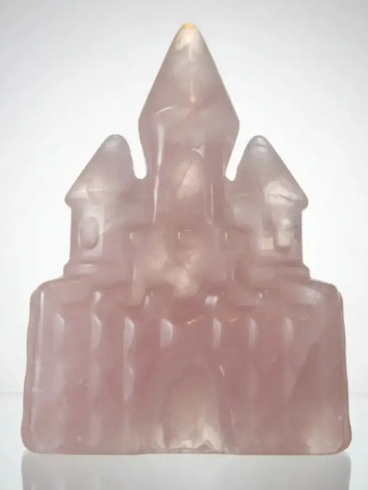 A translucent pale pink carved castle figurine, likely made of rose quartz or a similar stone, shown against a plain light gray background.