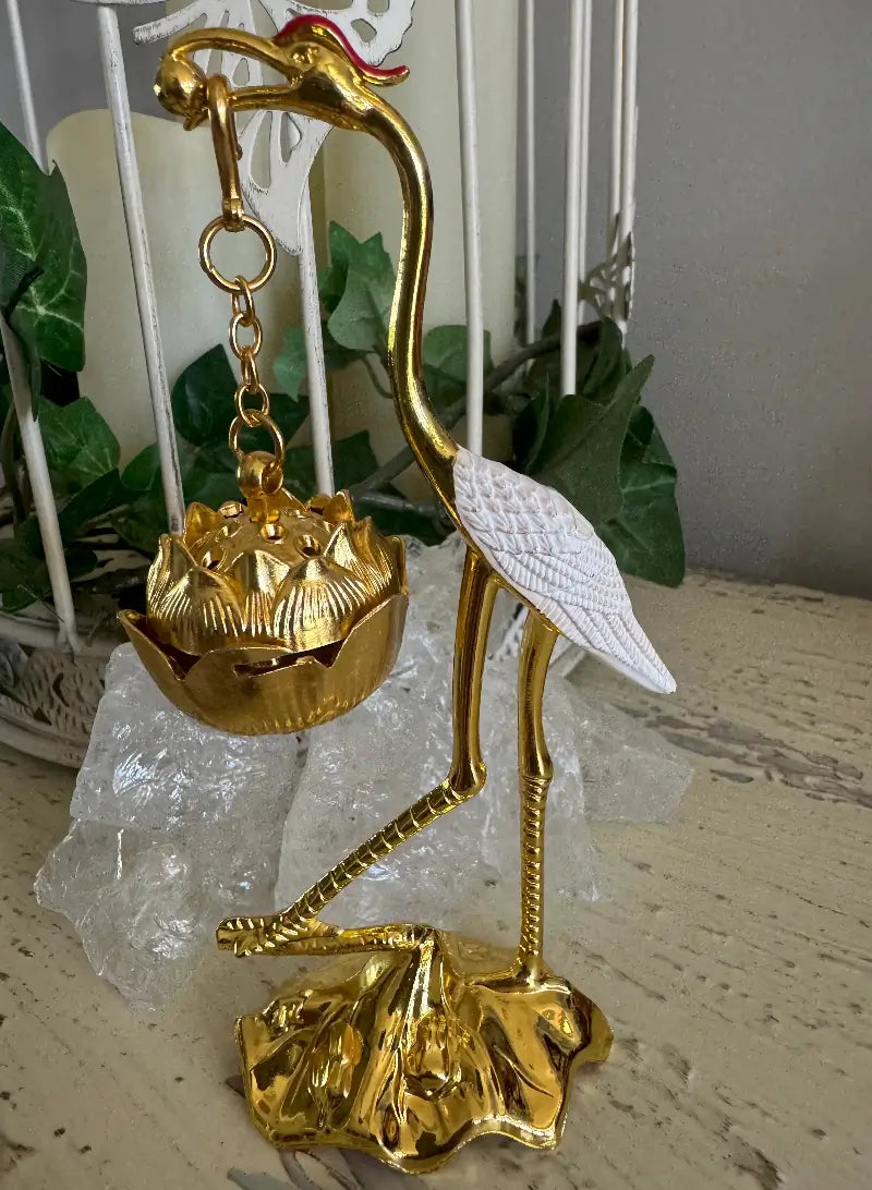 A decorative golden crane figurine holding a lotus-shaped hanging ornament, with a white textured body and base details, displayed on a tabletop with clear crystal blocks and green plants in the background.