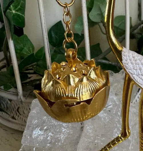 A gold-toned metal lotus pendant or ornament hanging from a chain, shown close up with decorative green leaves and white crystal stones in the background.