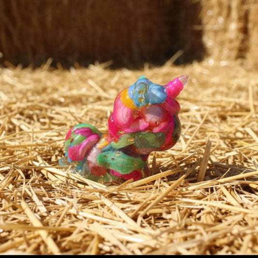 A little resin unicorn filled with carnival colors bright blue, yellow, pink, green dyed agate or stone sitting on a bed of straw. Product has gold toned metal shavings - orgonite like 