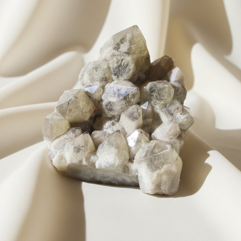 A cluster of natural crystals with rough, faceted shapes in pale beige, cream, and translucent gray tones, photographed on a soft fabric background