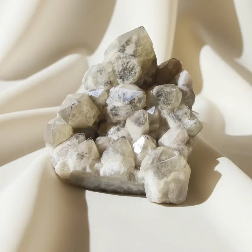 A cluster of natural crystals with rough, faceted shapes in pale beige, cream, and translucent gray tones, photographed on a soft fabric background