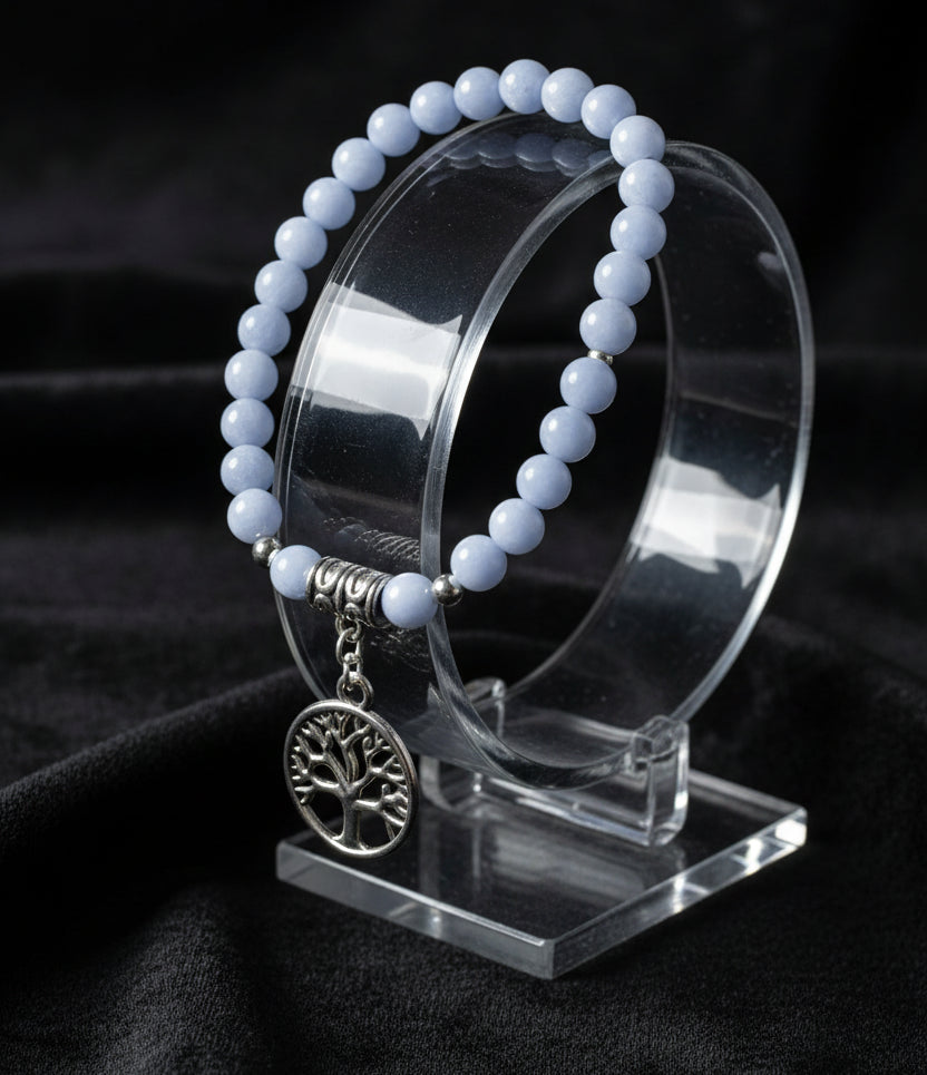 A beaded stretch bracelet featuring Angelite Crystal blue-gray round beads, a textured silver spacer bead, and a silver tree of life pendant charm, displayed on a black surface.